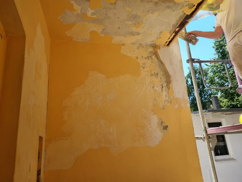 Ein Handwerker spachtelt die beschädigte Balkonecke, Wand und Decke zeigen noch alten abgeplatzten Putz