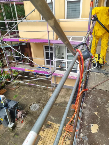 Ein Handwerker im gelben Schutzanzug reinigt die Fassade von oben mit dem Hochdruckreiniger, Blick auf den Hof darunter