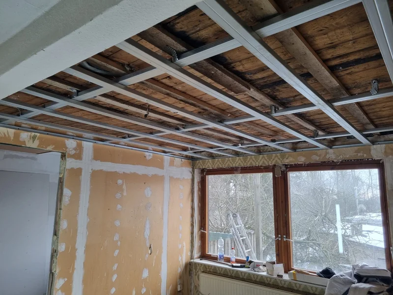 Fertig montierte Metallprofile an der Decke über orange grundierten Wänden, mit einer kleinen Leiter und Wasserflaschen auf der Fensterbank und schneebedeckten Bäumen im Hintergrund