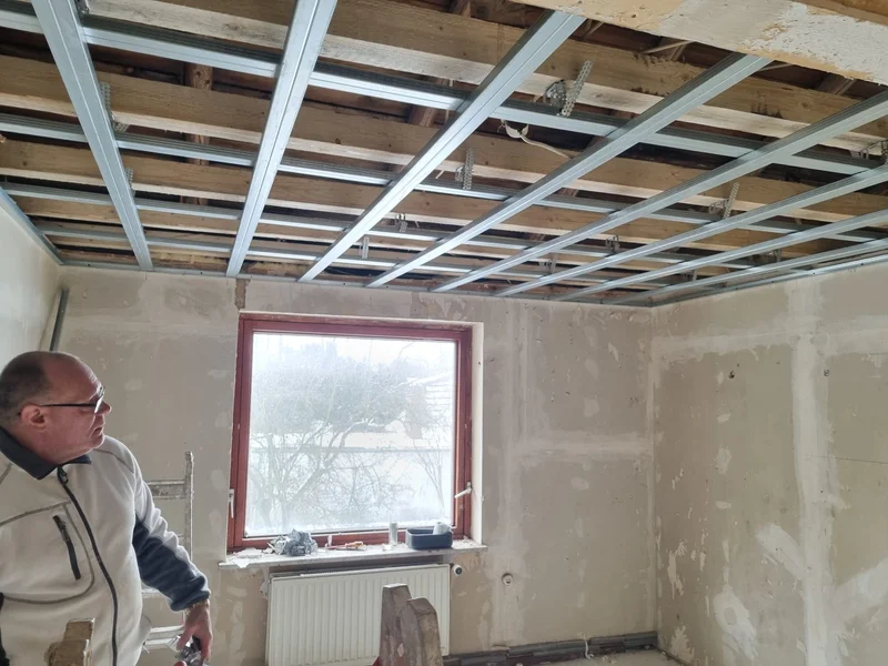 Nahaufnahme der fertigen Metalldeckenkonstruktion, ein Handwerker blickt nach oben in einem Raum mit grau verputzten Wänden und Blick auf Bäume durchs Fenster