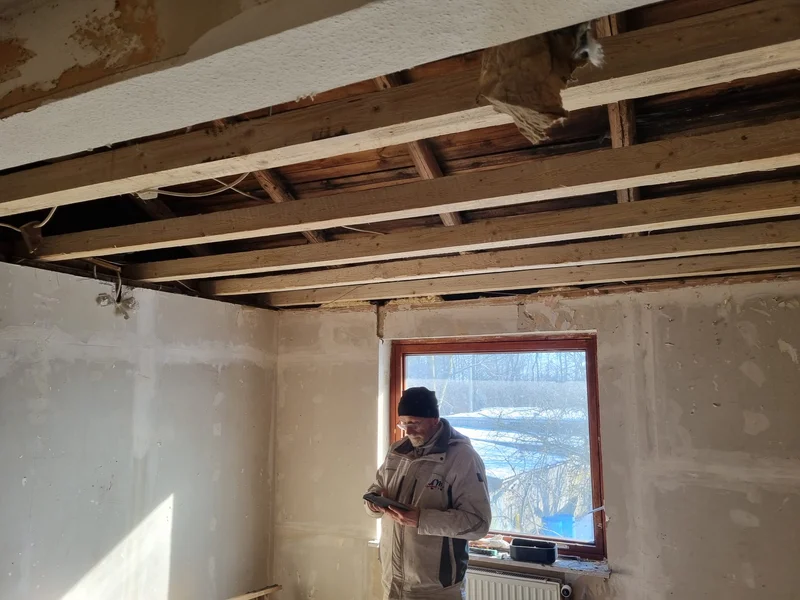 Die ursprünglichen Holzbalken der Decke vor dem Einbau der neuen Unterkonstruktion, ein Handwerker schaut am Fenster auf sein Tablet und draußen liegt eine verschneite Wiese
