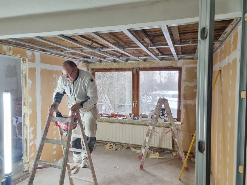 Übersicht des Raums mit fertig montierter Deckenkonstruktion über orange grundierten Wänden, ein Handwerker steht zwischen zwei Leitern am Fenster und draußen liegt Schnee
