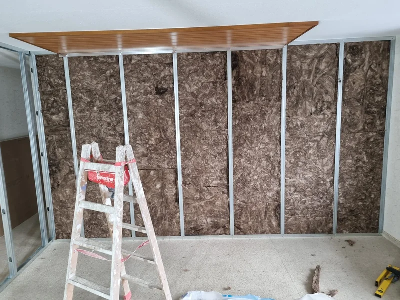 Mineralwolle zwischen den Metallständerprofilen der Wand, oben ein Streifen Holzverkleidung an der Decke und eine Leiter davor