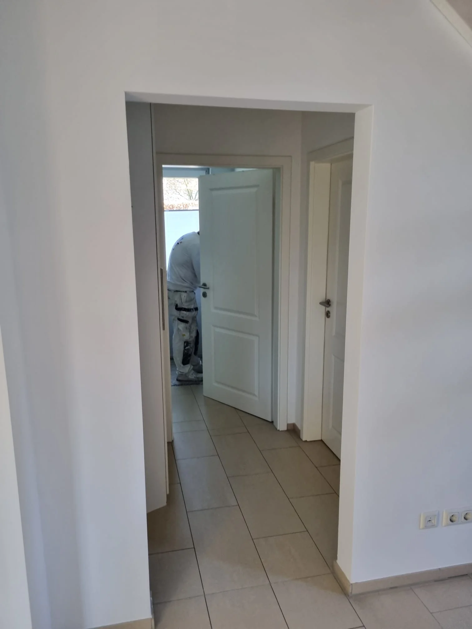 Blick durch einen frisch gestrichenen Flur mit weißen Türen und hellem Fliesenboden, im Hintergrund steht ein Handwerker in Malerkleidung an einer offenen Tür